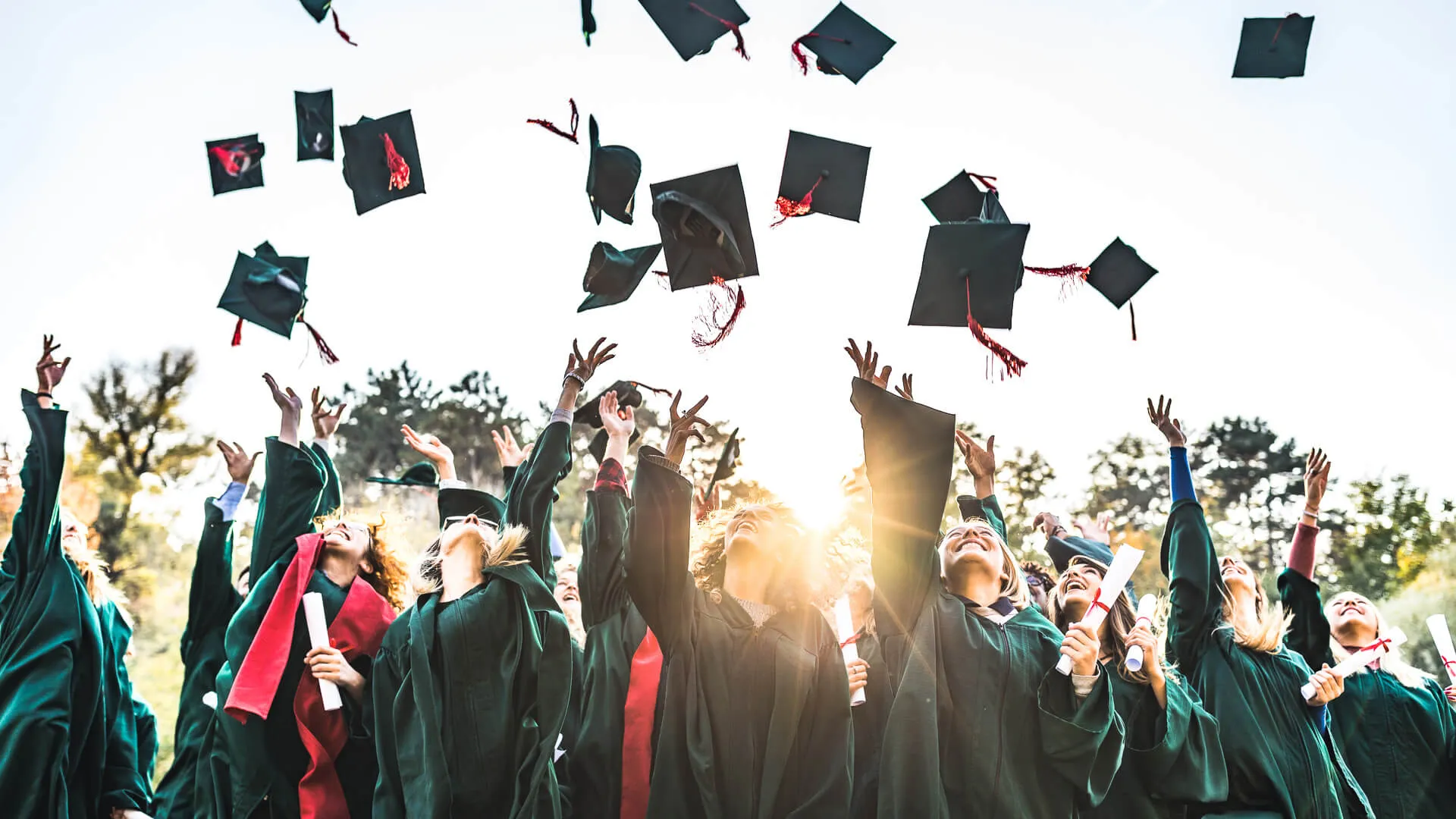 Large group of happy college students celebrating their graduation day outdoors while throwing their caps up in the air.